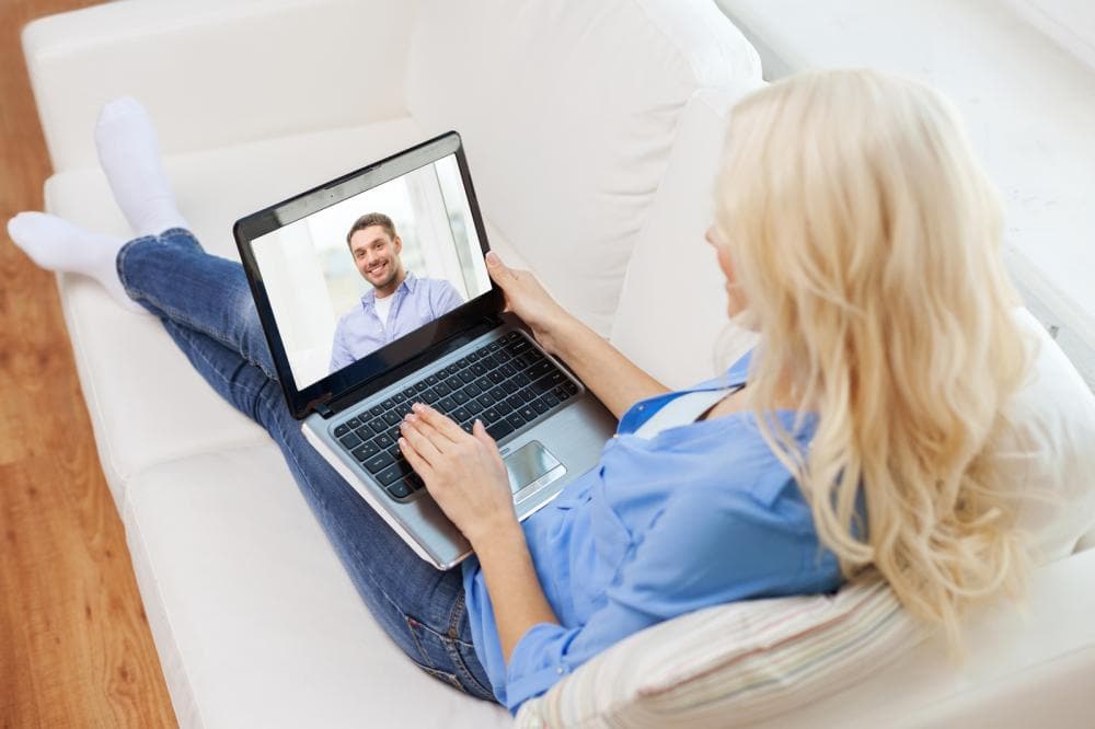 A blond woman is having a videochat. A computer sits on her lap and a man is on the computer, talking to her.